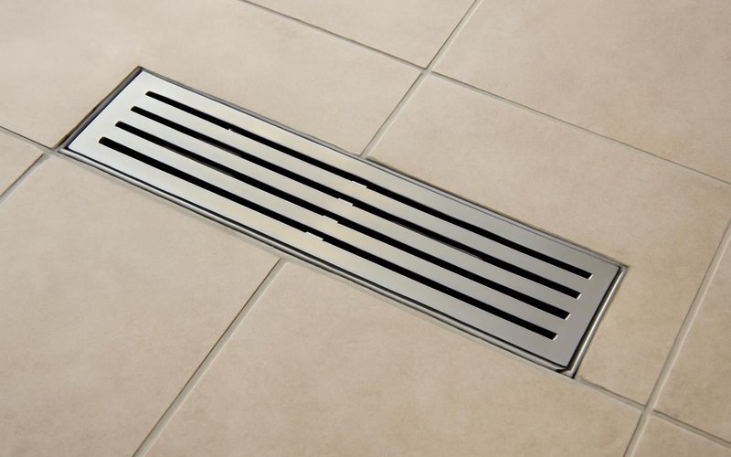 AI concept: An unbranded stainless steel linear shower drain with simple parallel slots set into beige tile, no writing